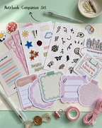 A notebook companion set flat-lay featuring various planner stickers, decorative labels, washi tape, a pen, and paper clips on a mint green background. The set includes pink, purple, and white elements with handwritten text like 'memories' and 'begin again'.