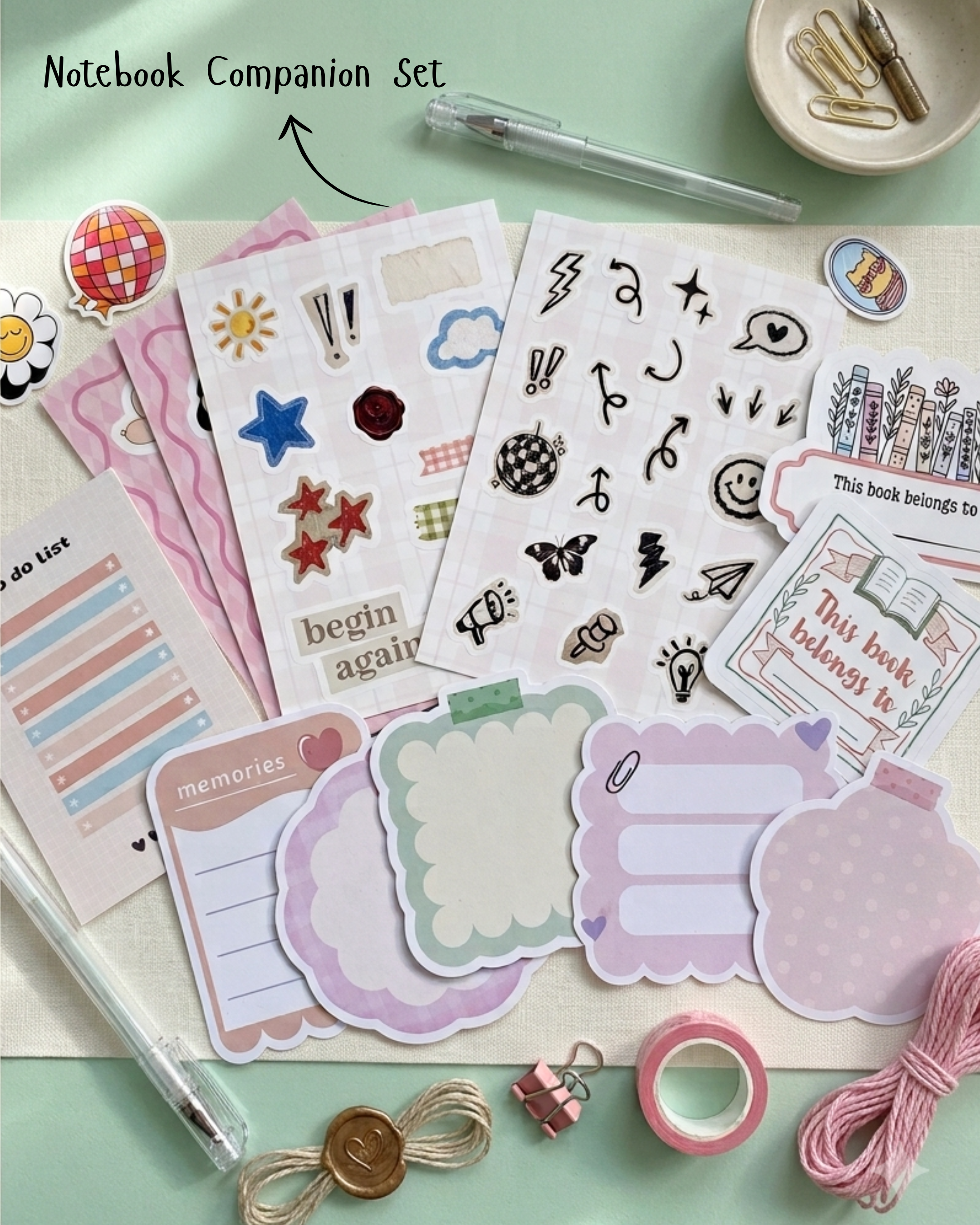 A notebook companion set flat-lay featuring various planner stickers, decorative labels, washi tape, a pen, and paper clips on a mint green background. The set includes pink, purple, and white elements with handwritten text like 'memories' and 'begin again'.